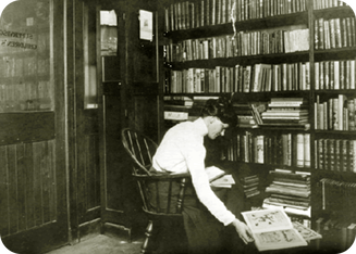 Anne Carroll Moore sitting in a room with a wall of books beside her